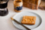 Slice of bread with peanut butter on a plate, knife beside it. Peanut butter jar and coffee maker in the background. Cozy kitchen scene.