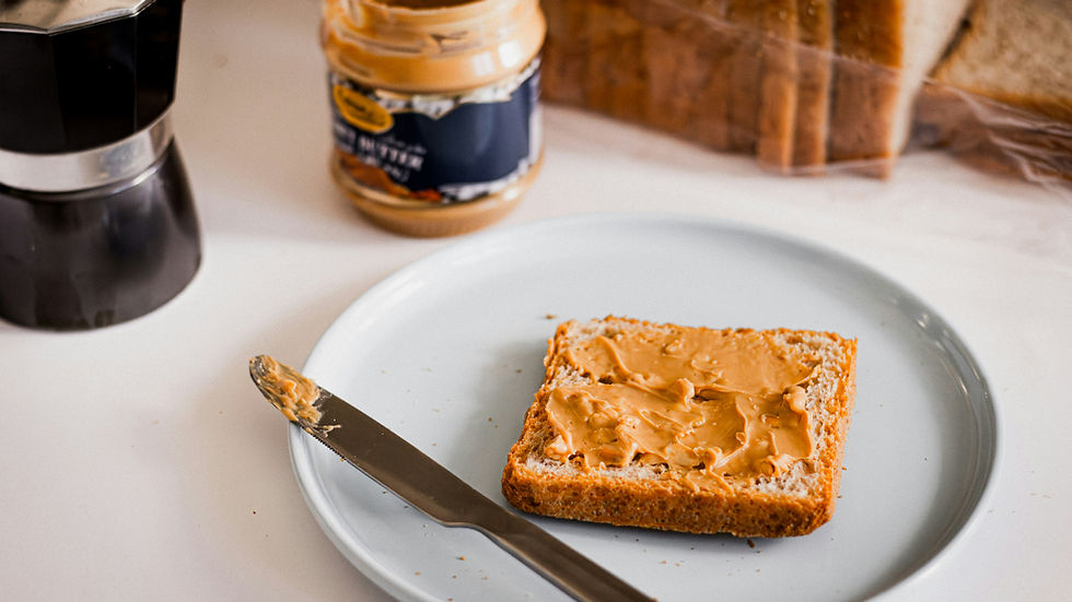 Slice of bread with peanut butter on a plate, knife beside it. Peanut butter jar and coffee maker in the background. Cozy kitchen scene.